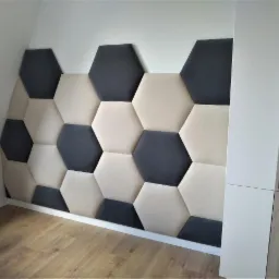 Panel ścienny z tapicerowanych heksagonów w odcieniach beżu i grafitu, tworzący geometryczny wzór w rogu pokoju obok białej szafki z drewnianymi elementami.