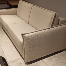 Jasna, dwuosobowa sofa z prostymi podłokietnikami i dwuczęściowym oparciem, ustawiona na marmurowej podłodze w minimalistycznym wnętrzu.