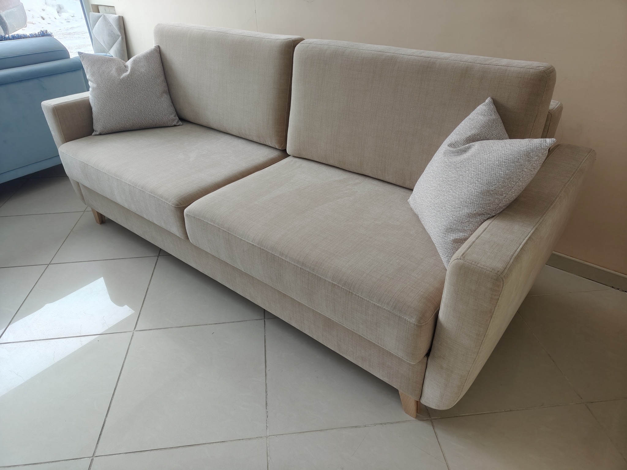 Beżowa sofa z poduszkami w salonie, widok z boku. Minimalistyczny design, jasne wnętrze, płytki podłogowe. Sofa na drewnianych nóżkach, komfortowy wygląd.