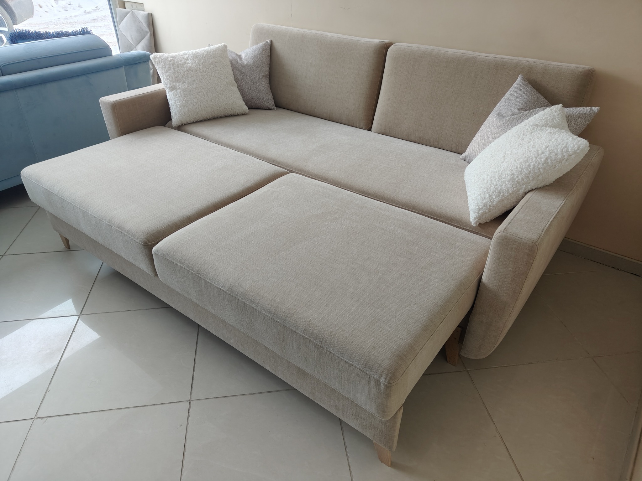 Beżowa sofa z funkcją spania i puszystymi poduszkami w salonie. Widoczny fragment innego mebla w tle. Jasne płytki na podłodze.