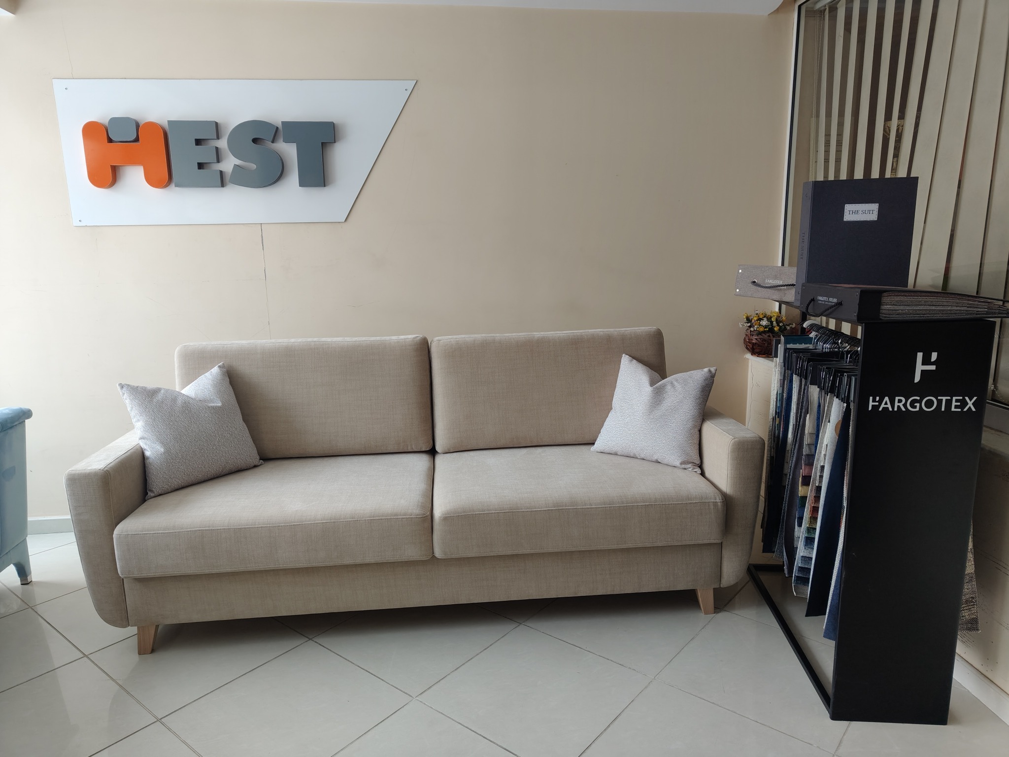 Beżowa sofa z poduszkami w salonie, obok ekspozytor z próbkami tkanin Hargotex. W tle logo firmy Hest na ścianie. Stonowana kolorystyka, naturalne światło.