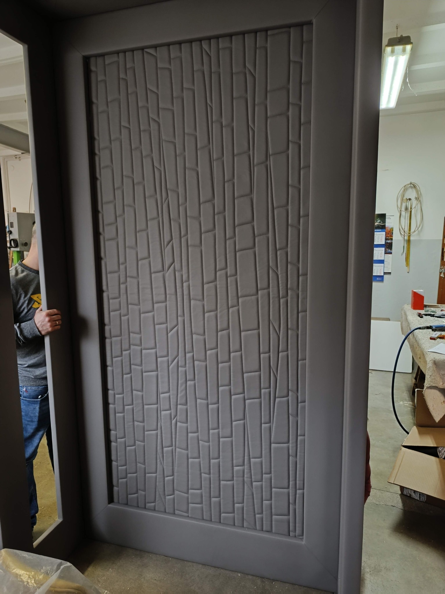 Szara, tapicerowana ściana akustyczna z geometrycznym wzorem, wykonana na zamówienie. Widoczny fragment osoby montującej. Oświetlenie warsztatowe w tle.