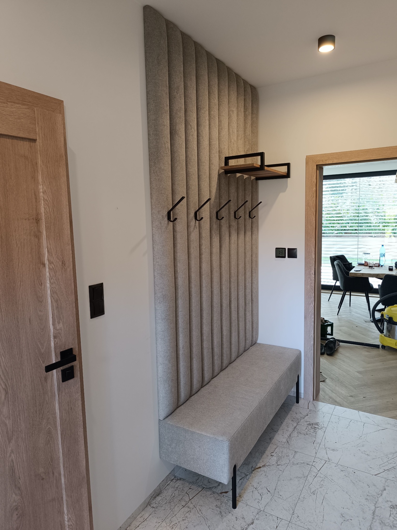 Tapicerowana ściana z siedziskiem i wieszakami w przedpokoju. Pionowe panele w beżowym odcieniu tworzą nowoczesny design. Na ścianie półka z drewnianymi elementami.