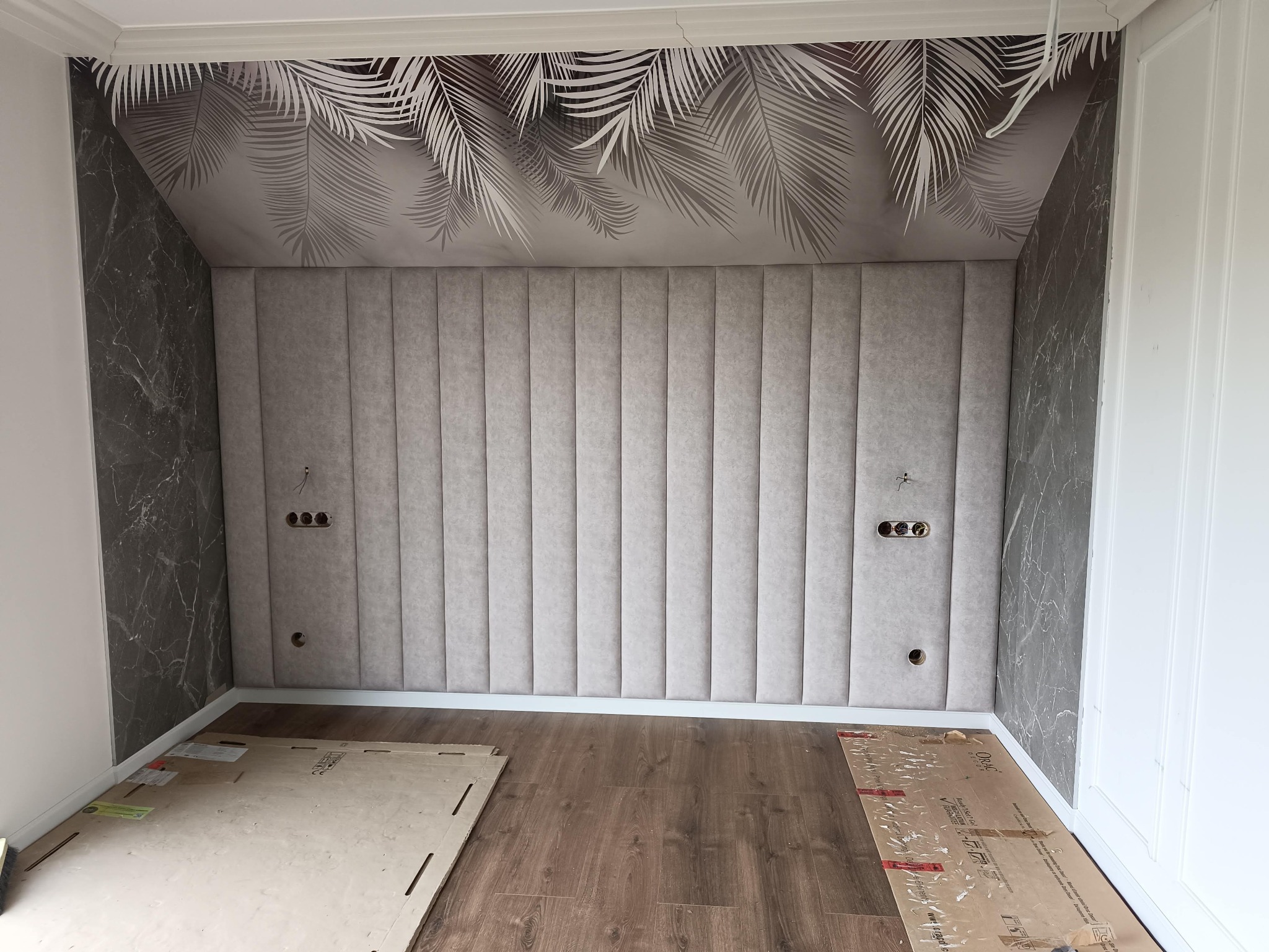 Tapicerowana ściana akustyczna z pionowymi panelami w odcieniach szarości, z widocznymi gniazdkami elektrycznymi, marmurową tapetą i motywem liści na suficie.