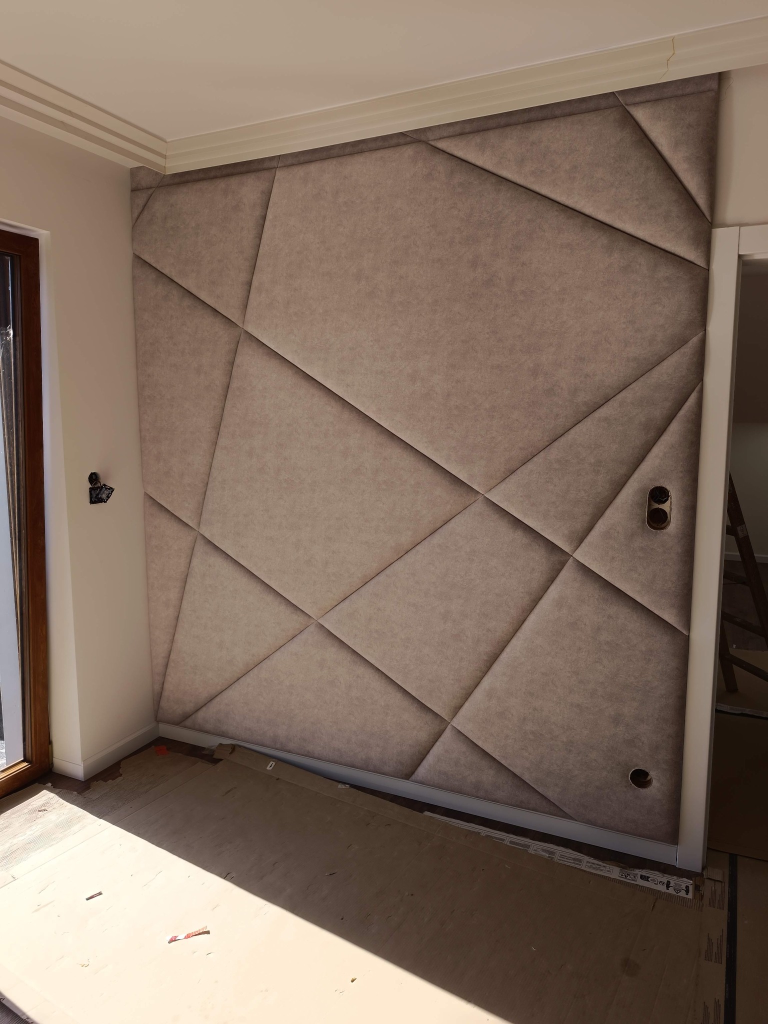 Tapicerowana ściana z geometrycznym wzorem w beżowym odcieniu, z widocznymi gniazdkami elektrycznymi i fragmentem okna. Podłoga zabezpieczona kartonem.