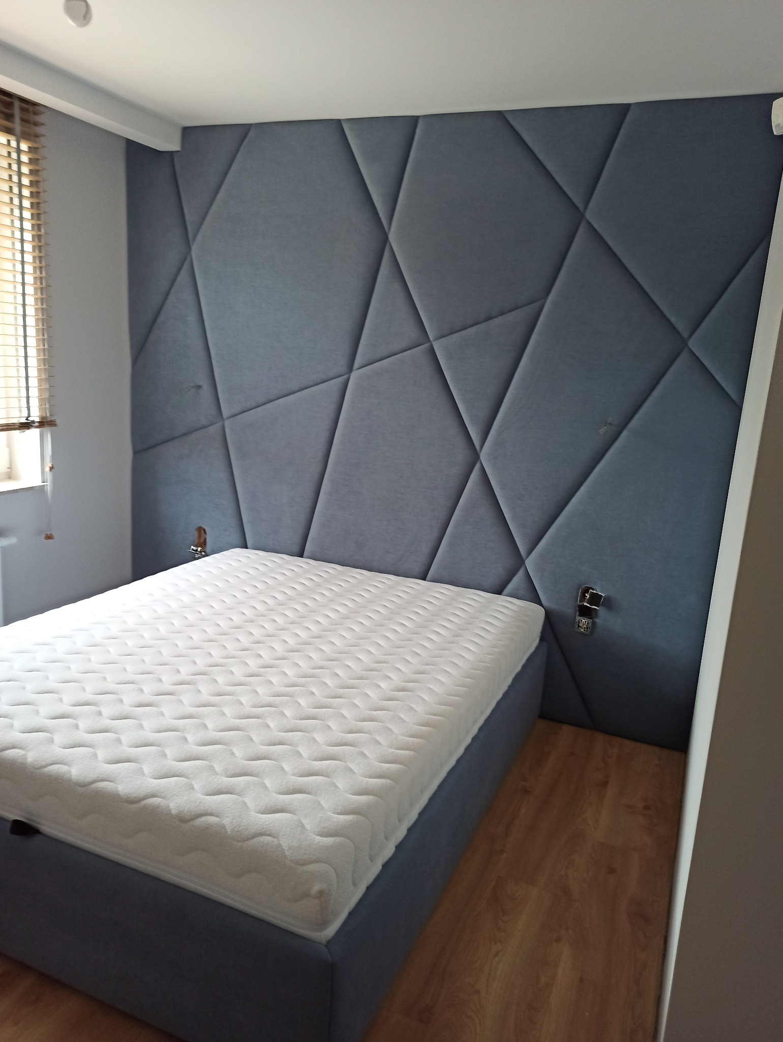 Sypialnia z tapicerowanym, geometrycznym zagłówkiem w odcieniach szarości. Nowoczesny design, biały materac, drewniane żaluzje w tle. Akustyczna aranżacja wnętrza.
