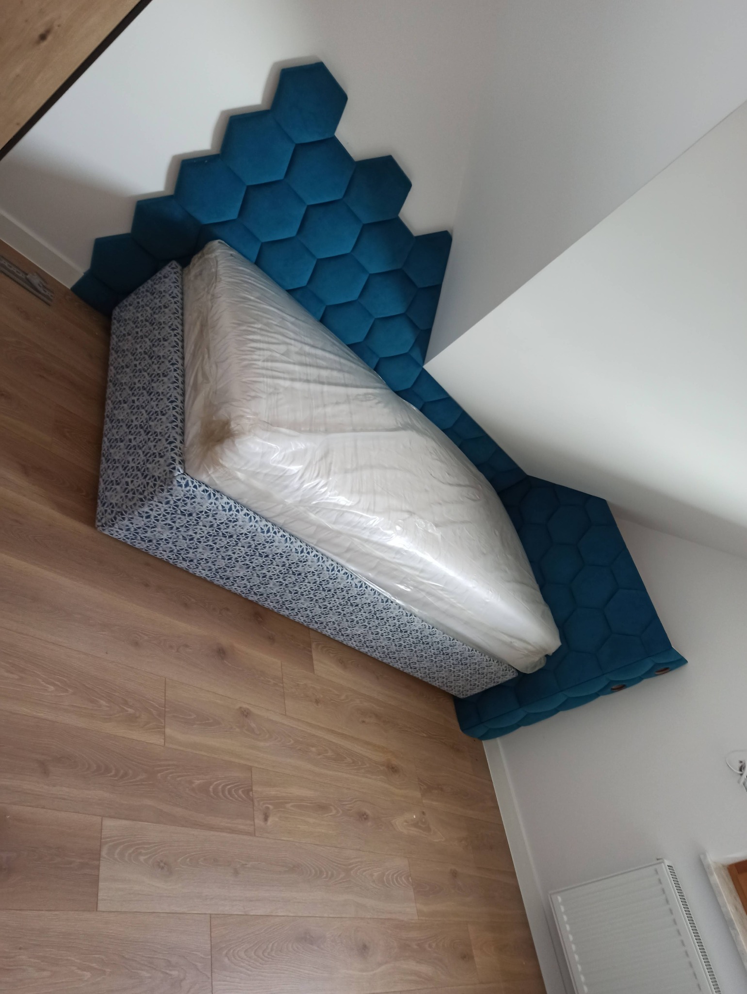 Tapicerowana sypialnia z wezgłowiem w kształcie plastra miodu, materac w folii. Geometryczny wzór na ramie łóżka, drewniana podłoga. Ujęcie z góry, ukośne.