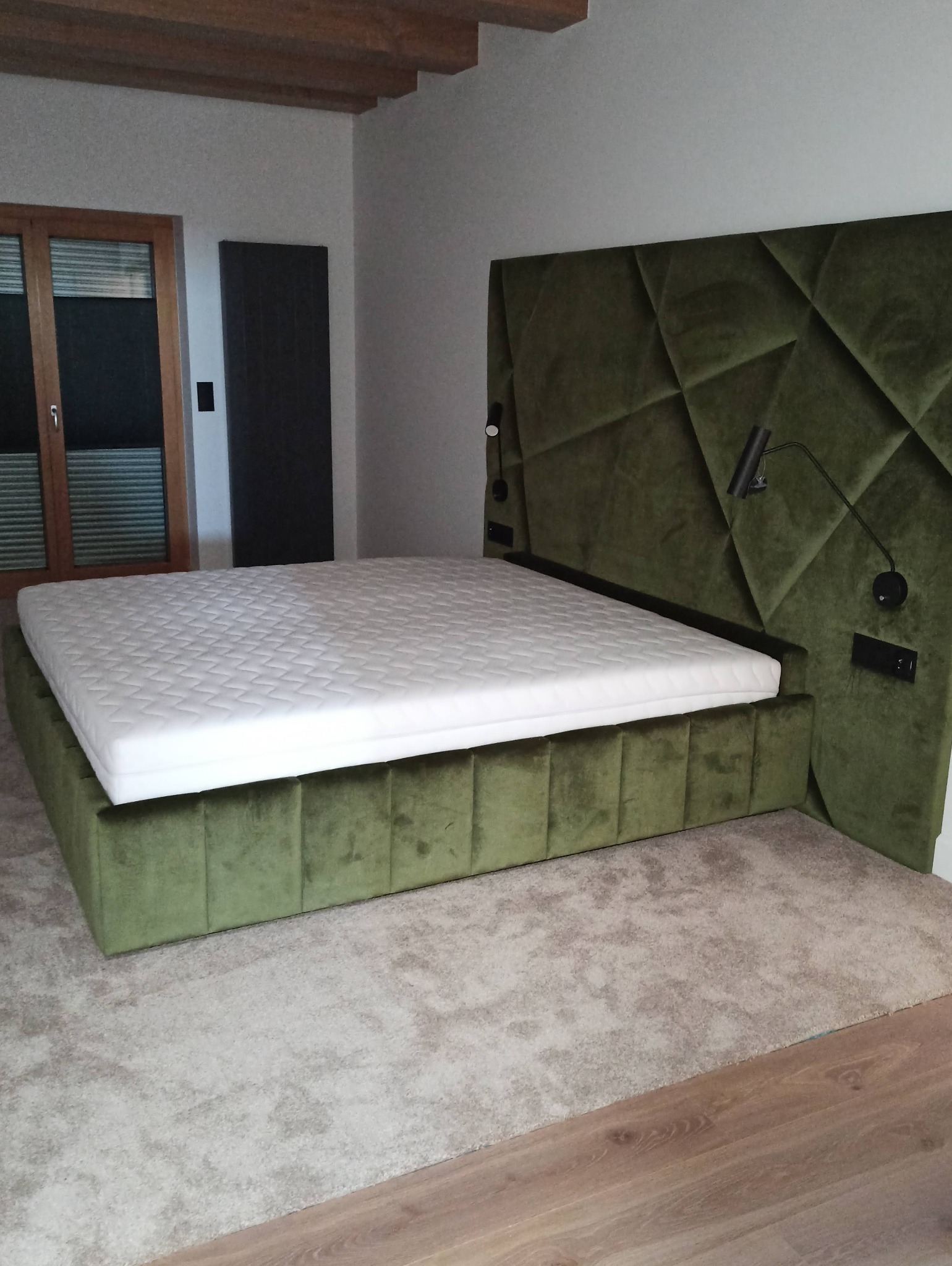 Nowoczesne łóżko tapicerowane w odcieniach zieleni z geometrycznym zagłówkiem i lampkami nocnymi, na tle jasnej sypialni z drewnianymi elementami.