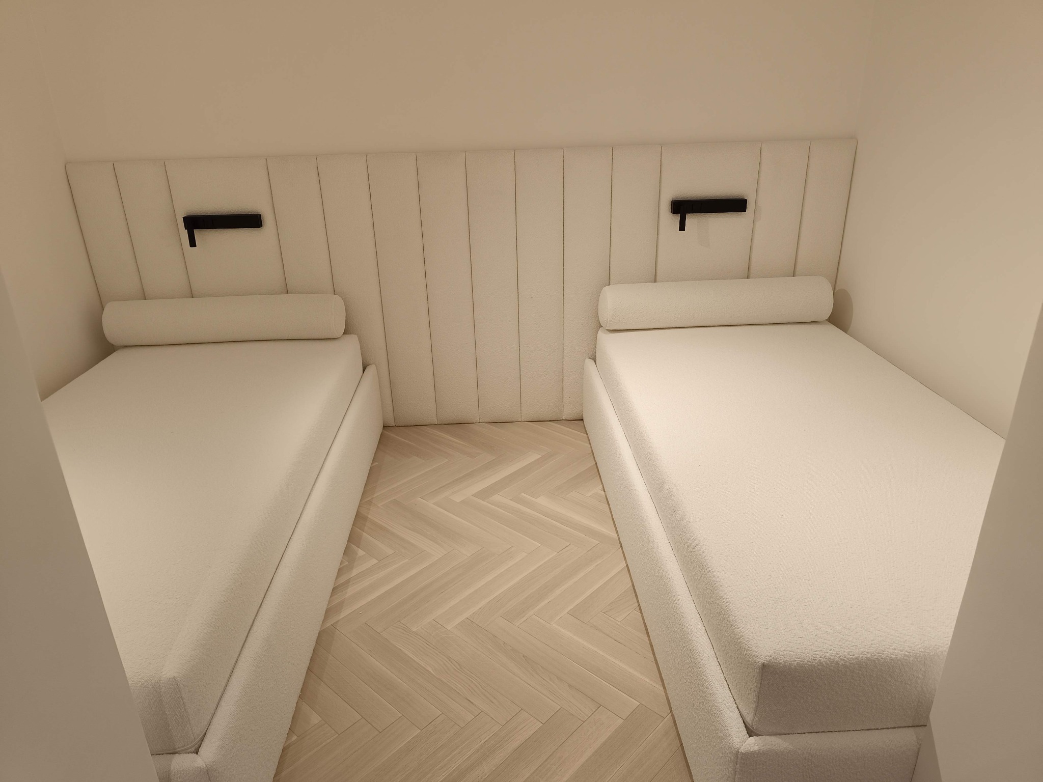 Dwa białe, tapicerowane łóżka pojedyncze z zagłówkami w wąskiej sypialni. Na ścianie czarne lampki. Parkiet ułożony w jodełkę. Minimalistyczny design.