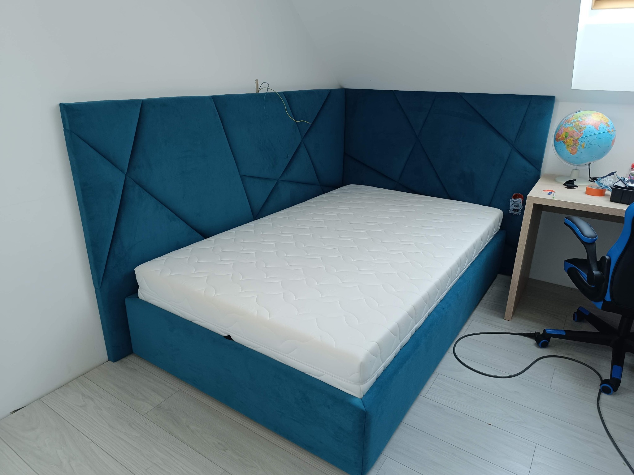 Niebieskie, tapicerowane łóżko z geometrycznym zagłówkiem w pokoju na poddaszu. Widoczne biurko z globusem i krzesło obok łóżka. Nowoczesny design.