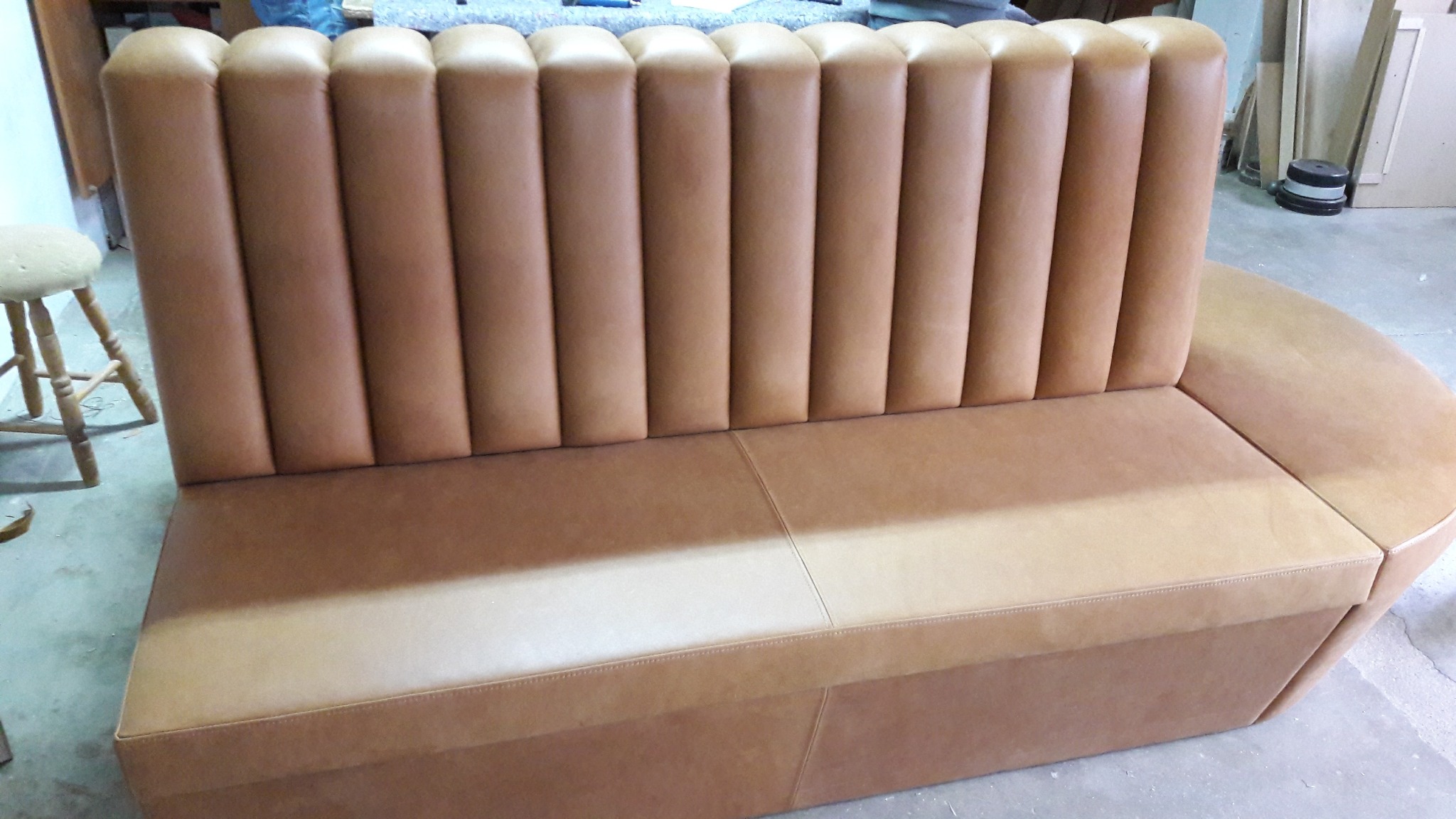 Tapicerowana sofa w odcieniach brązu z pionowymi przeszyciami na oparciu, przeznaczona do restauracji lub baru. Widoczny fragment krzesła barowego.