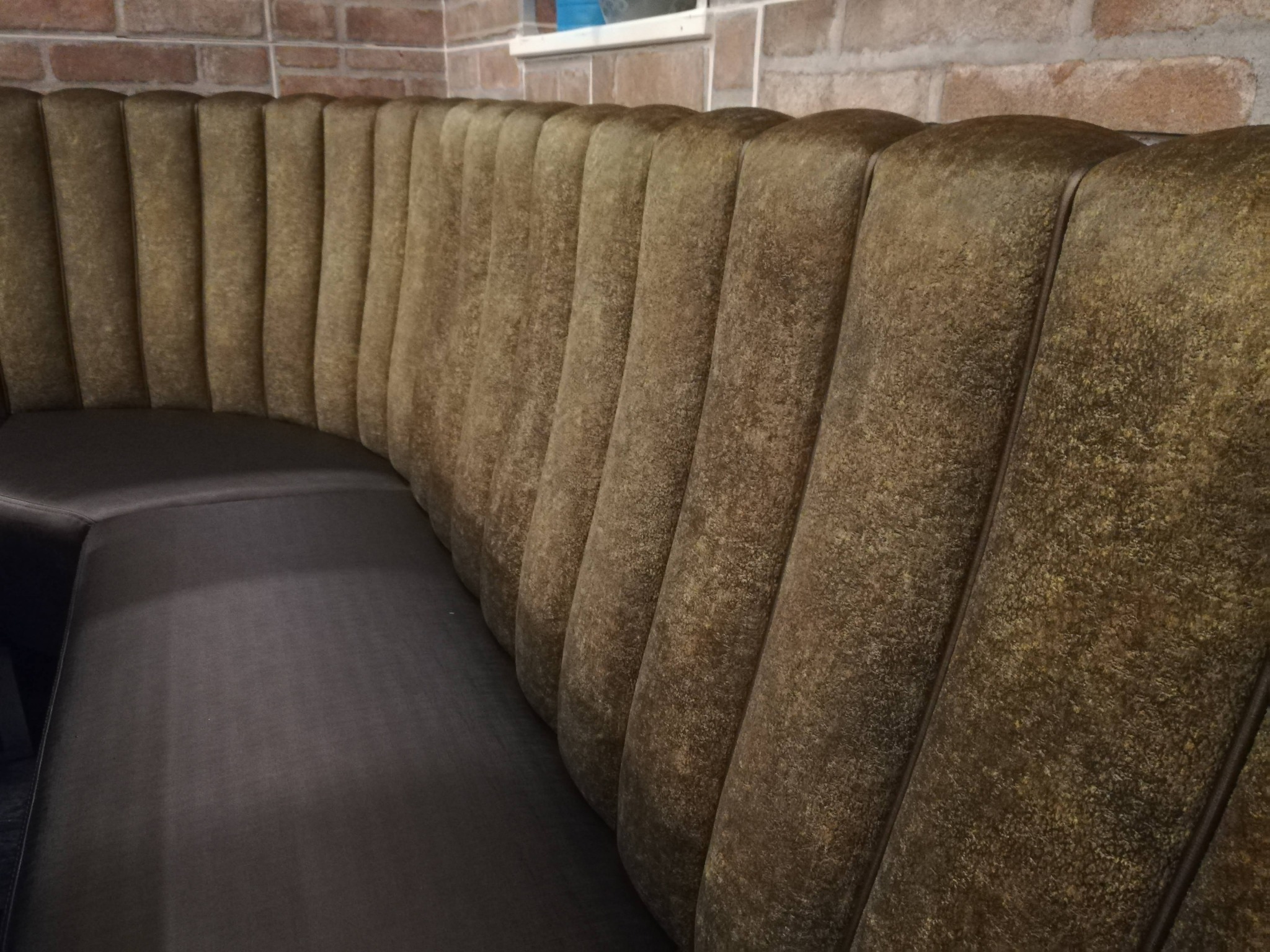 Zakrzywiona, tapicerowana sofa w odcieniach brązu, z pionowymi przeszyciami, na tle ceglanej ściany. Widok z boku, ukazujący detale faktury materiału.