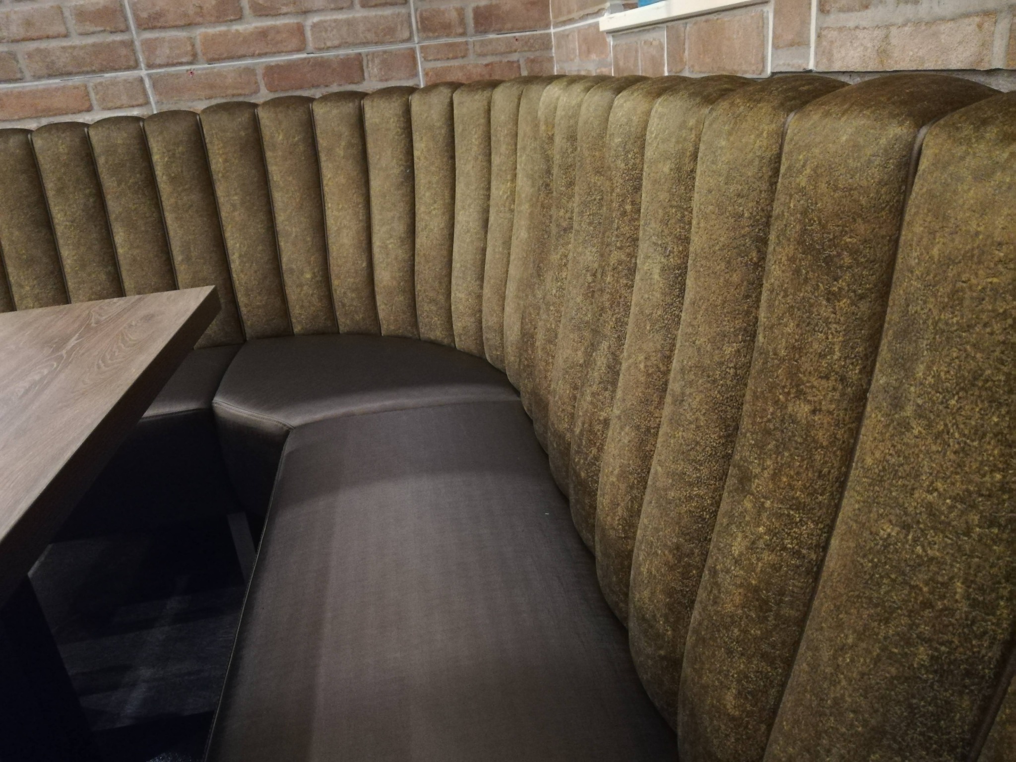 Tapicerowana, półokrągła sofa w odcieniach brązu, z drewnianym stolikiem, na tle ceglanej ściany. Widok z boku, ukazujący detale tapicerki.