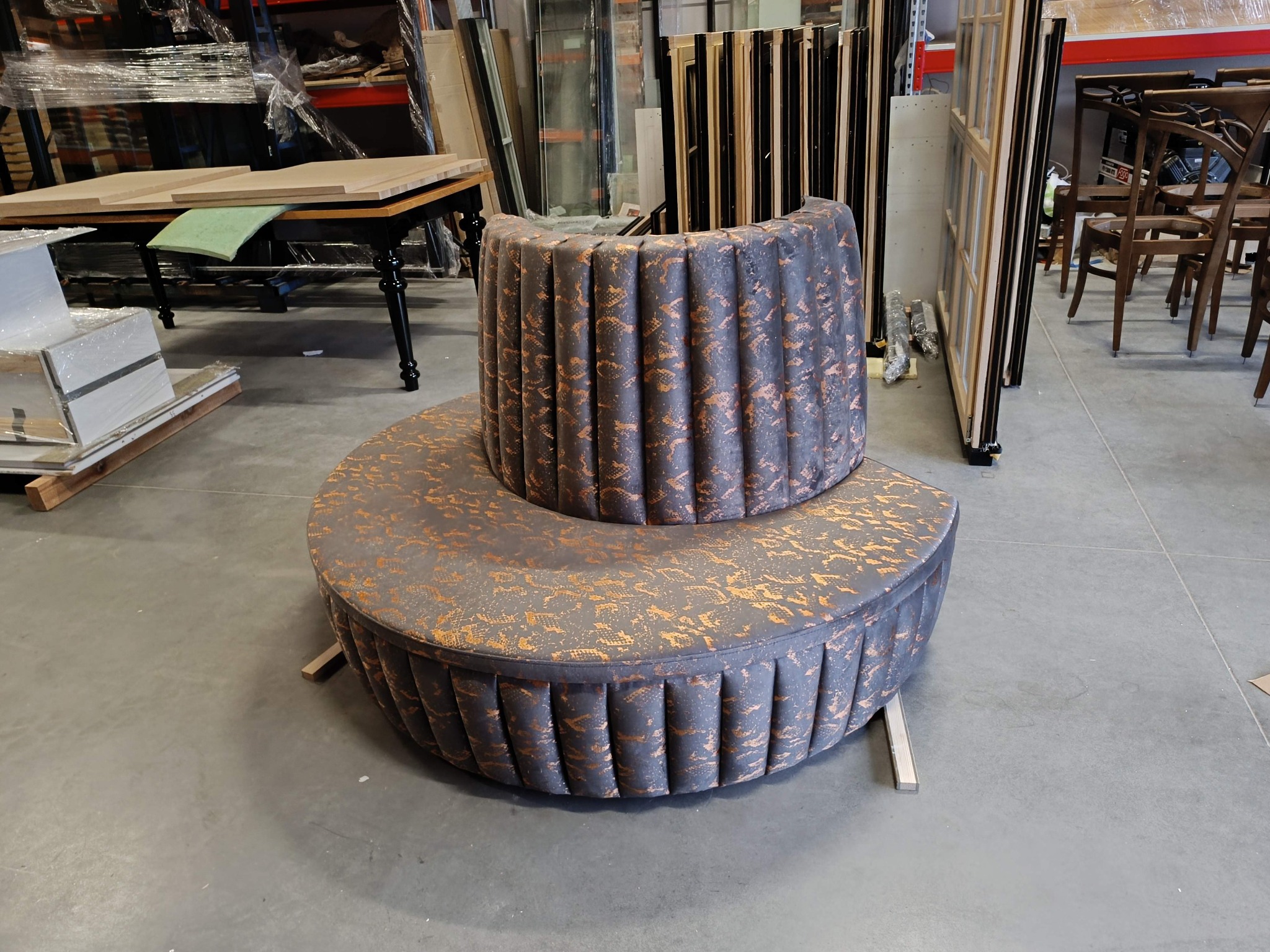 Okrągła, dwupoziomowa pufa tapicerowana w odcieniach szarości i brązu, z dekoracyjnym wzorem. Widoczne elementy warsztatu w tle.