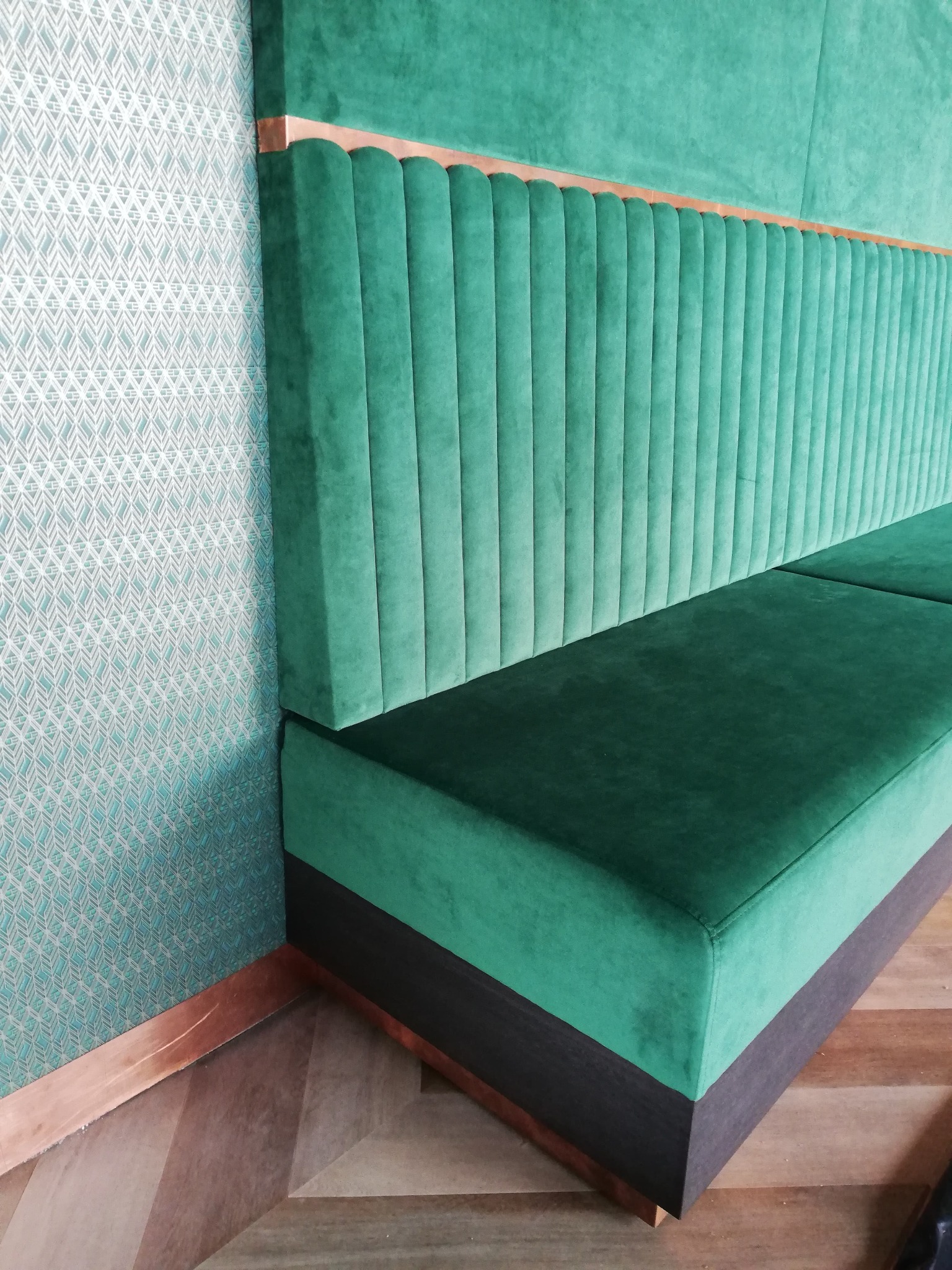 Szmaragdowa sofa tapicerowana z pionowymi przeszyciami, drewnianym akcentem i wzorzystą tapetą w tle. Ujęcie z perspektywy narożnika, podkreślające detale wykończenia.