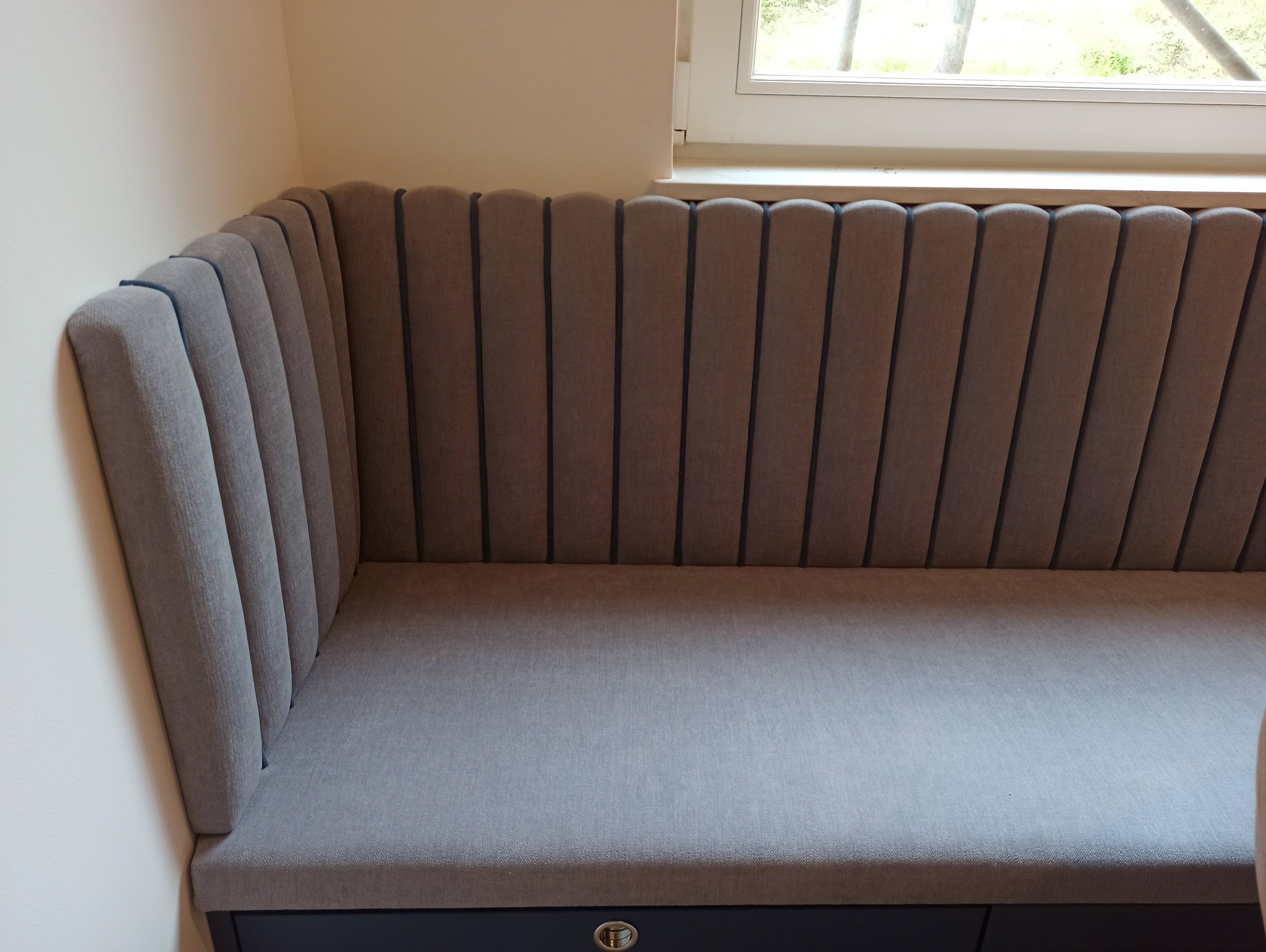 Tapicerowany narożnik wnękowy w odcieniach szarości, z pionowymi przeszyciami na oparciu, umieszczony przy oknie. Widoczny fragment siedziska i oparcia.