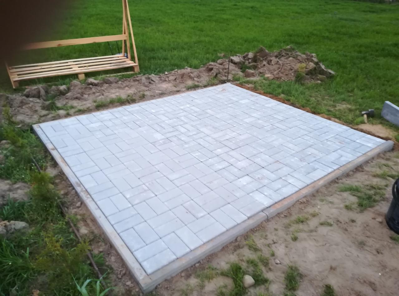 Świeżo ułożona, szara kostka brukowa w jodełkę na przygotowanym podłożu, widoczne obrzeża, w tle trawnik i drewniana konstrukcja.