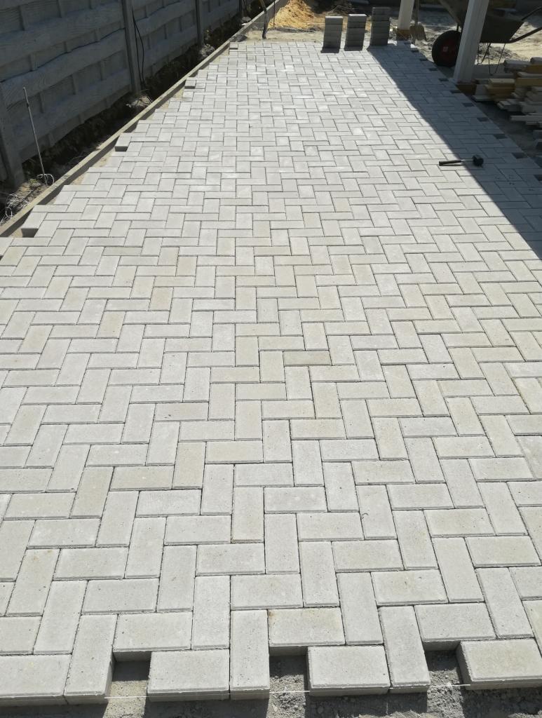 Ułożona szara kostka brukowa w jodełkę, widoczne niedokończone obrzeża i narzędzia w tle.