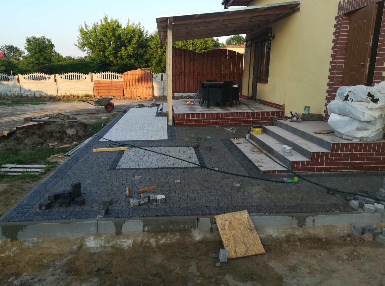 Częściowo ułożona nawierzchnia z kostki brukowej w dwóch kolorach, obok domu z tarasem i schodami, widoczne narzędzia i materiały budowlane.