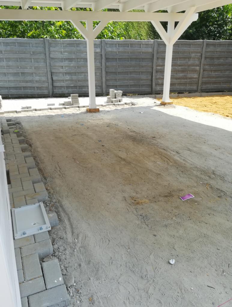 Etap przygotowania podłoża pod nawierzchnię z kostki brukowej w Krotoszynie, widoczne ułożone obrzeża i stosy kostki w tle, całość pod zadaszeniem.