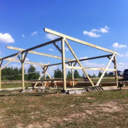 Wznoszona konstrukcja drewniana na betonowych fundamentach, widoczne elementy szkieletu budynku, w tle las i błękitne niebo z chmurami, obok zaparkowany samochód.