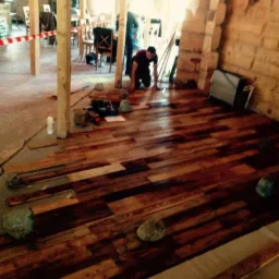 Montaż drewnianej podłogi z wykorzystaniem nieregularnych desek w surowym wnętrzu, z widocznymi narzędziami i obciążnikami z kamieni.