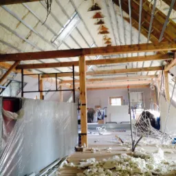 Poddasze w trakcie ocieplania pianką PUR, widoczne drewniane belki konstrukcyjne, fragment ściany zabezpieczony folią i resztki pianki na podłodze.