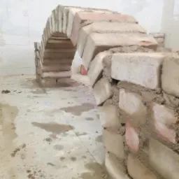 Wznoszenie łukowatego przejścia z cegieł, widoczne ślady zaprawy na podłodze, surowy stan wykończenia wnętrza.