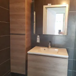 Nowoczesna łazienka z drewnopodobną szafką pod umywalką, lustrem z oświetleniem i wysoką szafką boczną, szare płytki na ścianach i podłodze, widoczne gniazdko elektryczne przed montażem.