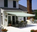 Zadaszony taras z markizą w biało-zielone pasy przy białym domu. Na tarasie stół i krzesła ogrodowe oraz donice z kwiatami.