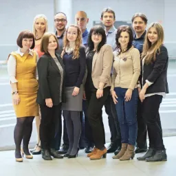 Grupa uśmiechniętych lektorów i lektorek w różnym wieku, ubranych w stylu biznesowym, stojących w jasnym, nowoczesnym wnętrzu biurowym z przeszklonymi balustradami.