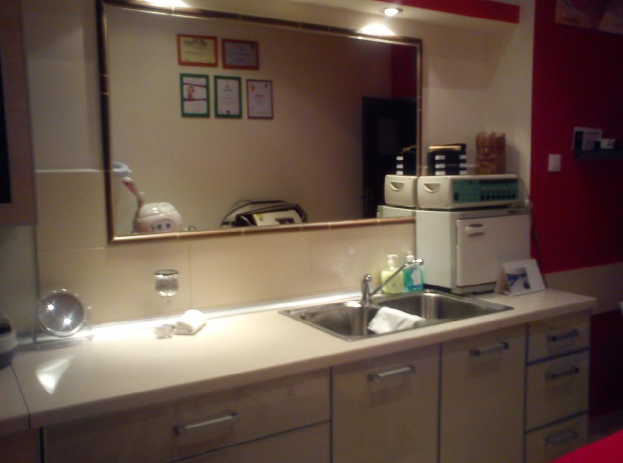 Wnętrze gabinetu z umywalką, sprzętem kosmetycznym i lustrem, w którym odbijają się certyfikaty na ścianie.