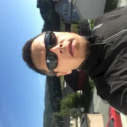 Autoportret mężczyzny w okularach przeciwsłonecznych na tle krajobrazu Święciechowa. Widoczne budynki i zieleń. Nietypowy kąt ujęcia.