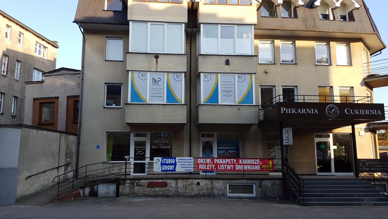 Fasada budynku z biurami projektowymi i piekarnią/cukiernią w słoneczny dzień, widoczne szyldy firm i okna z reklamami usług projektowych.