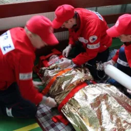 Trzech ratowników medycznych w czerwonych uniformach udziela pomocy osobie leżącej na noszach, przykrytej folią termiczną i z maską tlenową; jeden z ratowników trzyma butlę z tlenem.