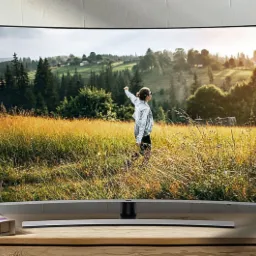 Zakrzywiony telewizor Samsung wyświetlający krajobraz z osobą w polu, ustawiony na drewnianej komodzie obok roślin doniczkowych i dekoracji.