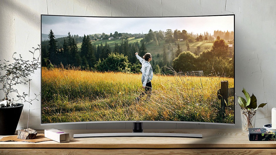 Zakrzywiony telewizor Samsung wyświetlający krajobraz z osobą w polu, ustawiony na drewnianej komodzie obok roślin doniczkowych i dekoracji.