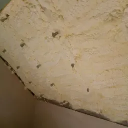 Nierównomiernie nałożona pianka izolacyjna PUR na poddaszu, widoczne metalowe kołki mocujące, fragment ściany pomalowanej na beżowo w dolnym rogu.