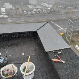 Fragment dachu pokrytego papą z obróbką blacharską, widoczne narzędzia dekarskie i pojemniki, w tle rozmyty krajobraz miejski w mglisty dzień.