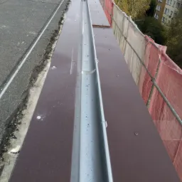 Nowy system rynnowy zamontowany na dachu z papy, widok z góry, zabezpieczony siatką rusztowaniową, w tle budynki mieszkalne.