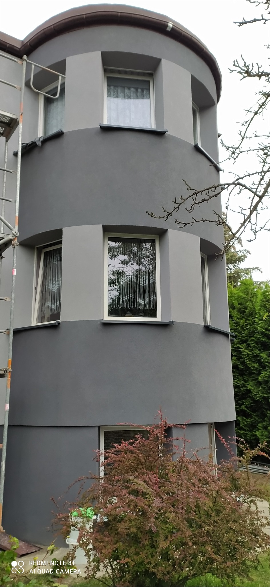 Dwukolorowa elewacja budynku z zaokrąglonym narożnikiem, widoczne okna z firankami, fragment rusztowania i ozdobny krzew.