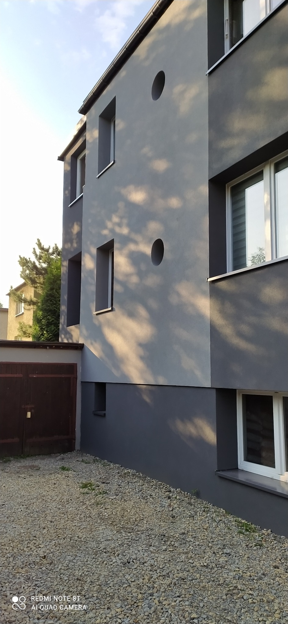 Dwukondygnacyjny budynek z szarą elewacją, okrągłymi otworami w ścianie i widocznymi cieniami drzew, z brązową bramą garażową i żwirowym podłożem.