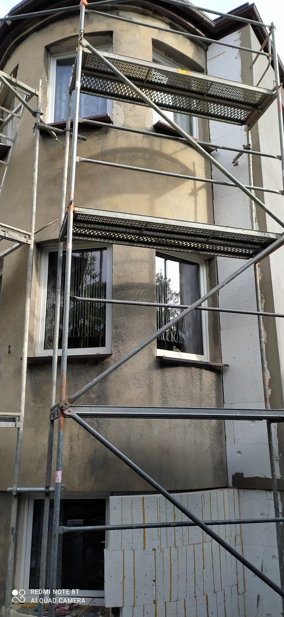 Budynek w trakcie prac elewacyjnych, widoczne rusztowania, fragment ocieplonej ściany z białym styropianem oraz fragment ściany przed ociepleniem z widocznymi oknami.