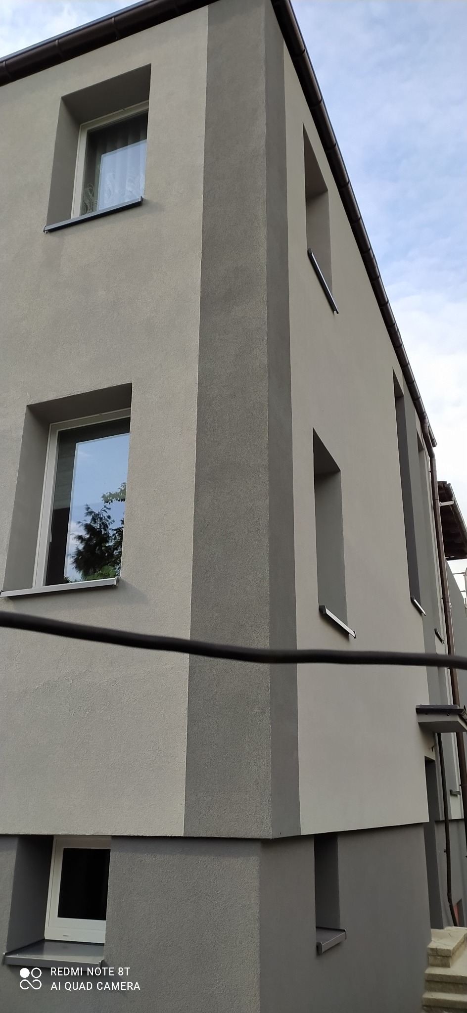 Elewacja budynku po remoncie, widoczne okna z odbiciem drzew, szara kolorystyka ścian z pionowymi pasami w odcieniach szarości.