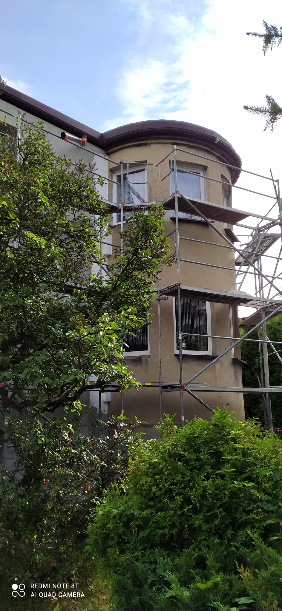 Fragment budynku z zaokrągloną częścią, na którym widać rusztowanie, a część elewacji jest w trakcie prac wykończeniowych; widok częściowo zasłonięty przez zieleń.