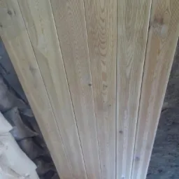 Ułożone pionowo deski z jasnego drewna o widocznych słojach, obok worki z materiałem budowlanym.