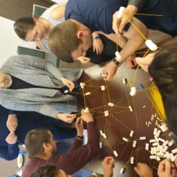Grupa osób pochylona nad stołem, budująca konstrukcję z makaronu spaghetti i pianek marshmallow, widok z góry.