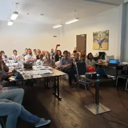 Sala konferencyjna podczas szkolenia: uczestnicy siedzą przy stołach z notatkami, prelegentka stoi obok laptopa i projektora, w tle wiszą obrazy z motywami ptak&oacute;w.