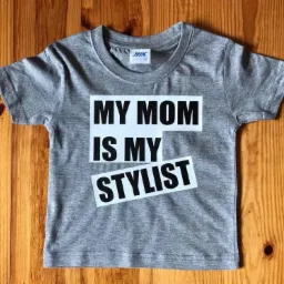 Szara koszulka dziecięca z nadrukiem 'MY MOM IS MY STYLIST' na drewnianym tle.
