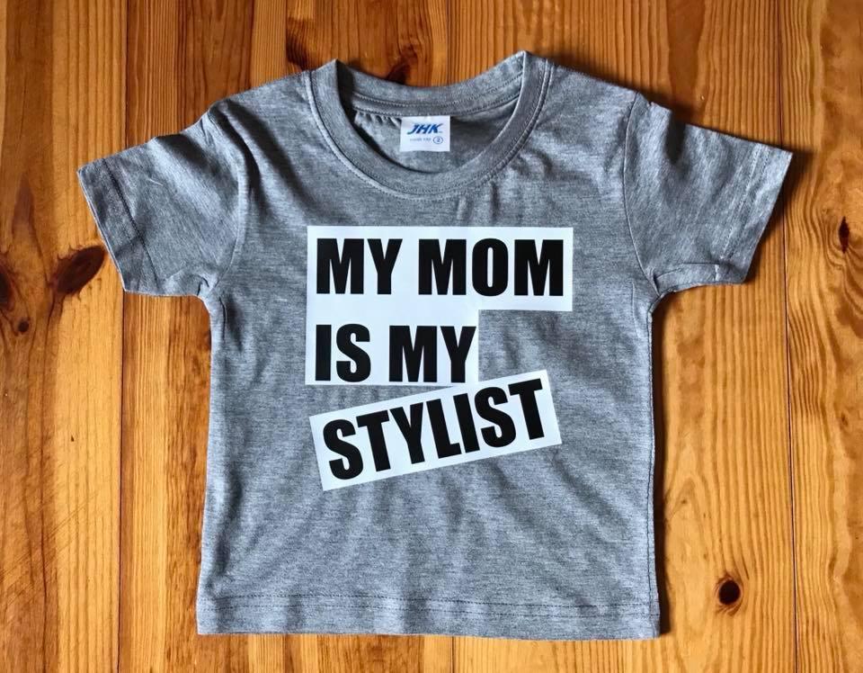 Szara koszulka dziecięca z nadrukiem 'MY MOM IS MY STYLIST' na drewnianym tle.